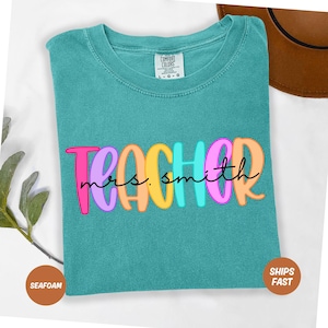 May include: Seafoam green Comfort Colors t-shirt with the word "TEACHER" in colourful bubble letters and "mrs. smith" in a cursive font. The shirt has a crew neck and short sleeves. A brown hat is visible in the background.