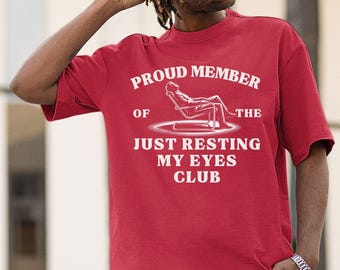 May include: A red t-shirt with the text "PROUD MEMBER OF THE JUST RESTING MY EYES CLUB" in white. The graphic shows a person reclining in a chair. The shirt is short-sleeved.