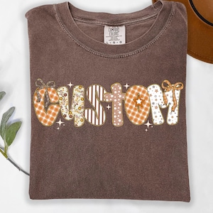 Puede incluir: Una camiseta marrón con la palabra "CUSTOM" en una fuente decorativa estampada. Las letras presentan diseños de cuadros, flores, rayas y estrellas en naranja, marrón y blanco. La camiseta tiene una etiqueta Comfort Colors.