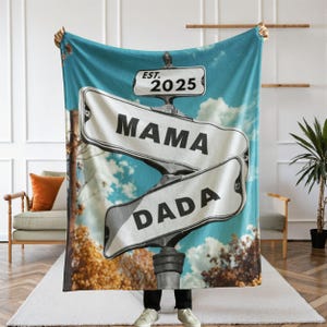 May include: A blue blanket featuring a road sign design with the words "MAMA" and "DADA" on separate signs. The sign also includes the text "EST. 2025" and is set against a sky and tree background.