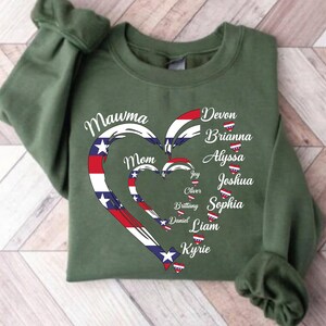 K&ouml;nnte beinhalten: Ein olivgr&uuml;nes Sweatshirt mit einem Herzdesign in Rot, Wei&szlig; und Blau, mit den Worten "Mawma Mom" und Namen wie Devon, Brianna und Alyssa. Das Design erinnert an die amerikanische Flagge.
