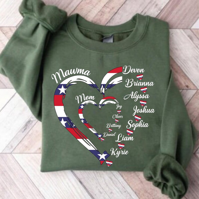 K&ouml;nnte beinhalten: Ein olivgr&uuml;nes Sweatshirt mit einem Herzdesign in Rot, Wei&szlig; und Blau, mit den Worten "Mawma Mom" und Namen wie Devon, Brianna und Alyssa. Das Design erinnert an die amerikanische Flagge.