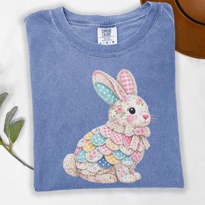 May include: A blue Comfort Colors t-shirt with a patchwork bunny design. The bunny is decorated with floral and polka dot patterns, a pink bow, and a fluffy tail. The shirt has a crew neck and short sleeves.