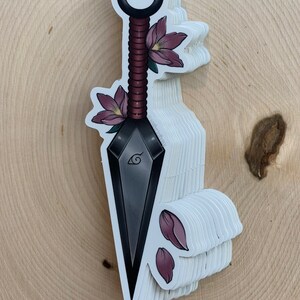 Puede incluir: Un kunai negro y gris con un mango rojo y blanco, decorado con flores rosas. El kunai tiene un dise&ntilde;o de espiral y un s&iacute;mbolo en la hoja.