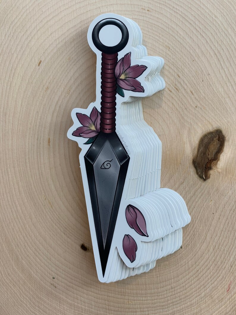 Puede incluir: Un kunai negro y gris con un mango rojo y blanco, decorado con flores rosas. El kunai tiene un dise&ntilde;o de espiral y un s&iacute;mbolo en la hoja.