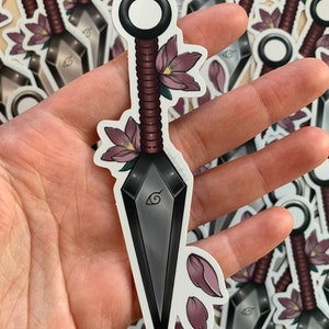 Puede incluir: Un kunai negro y plateado con un mango rojo y negro, decorado con flores y p&eacute;talos rosas. El kunai tiene un dise&ntilde;o de espiral en la hoja y un anillo circular en el mango.