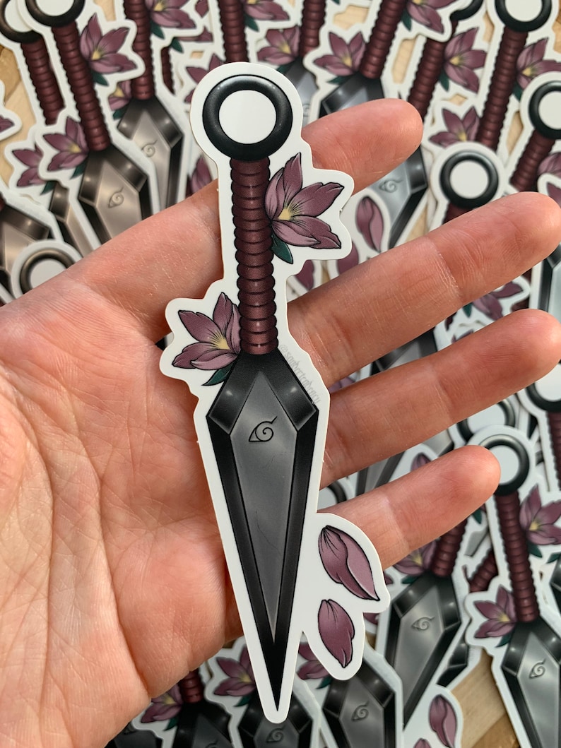 Puede incluir: Un kunai negro y plateado con un mango rojo y negro, decorado con flores y p&eacute;talos rosas. El kunai tiene un dise&ntilde;o de espiral en la hoja y un anillo circular en el mango.