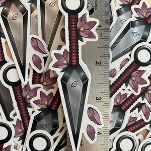 Puede incluir: Un conjunto de pegatinas que representan un cuchillo de lanzamiento estilizado con un mango de cuerda roja y flores rosas. El cuchillo tiene una hoja plateada con un s&iacute;mbolo negro. Las pegatinas est&aacute;n dispuestas alrededor de una regla, mostrando que miden aproximadamente 5 cm.