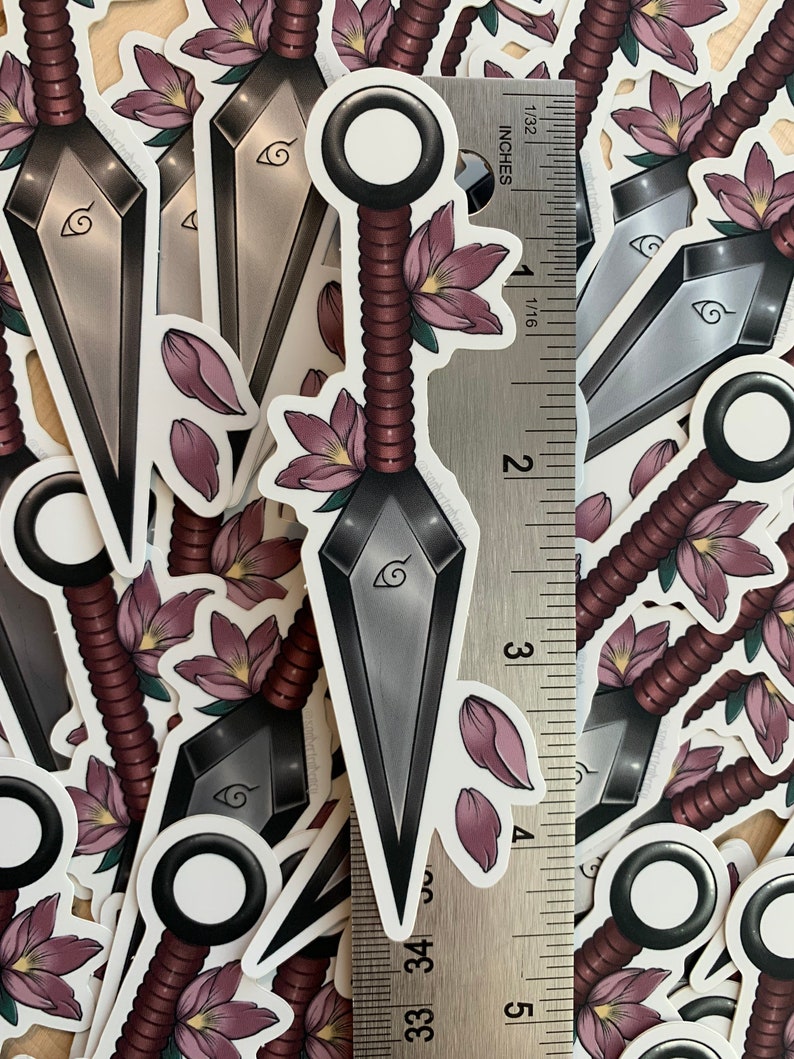 Puede incluir: Un conjunto de pegatinas que representan un cuchillo de lanzamiento estilizado con un mango de cuerda roja y flores rosas. El cuchillo tiene una hoja plateada con un s&iacute;mbolo negro. Las pegatinas est&aacute;n dispuestas alrededor de una regla, mostrando que miden aproximadamente 5 cm.