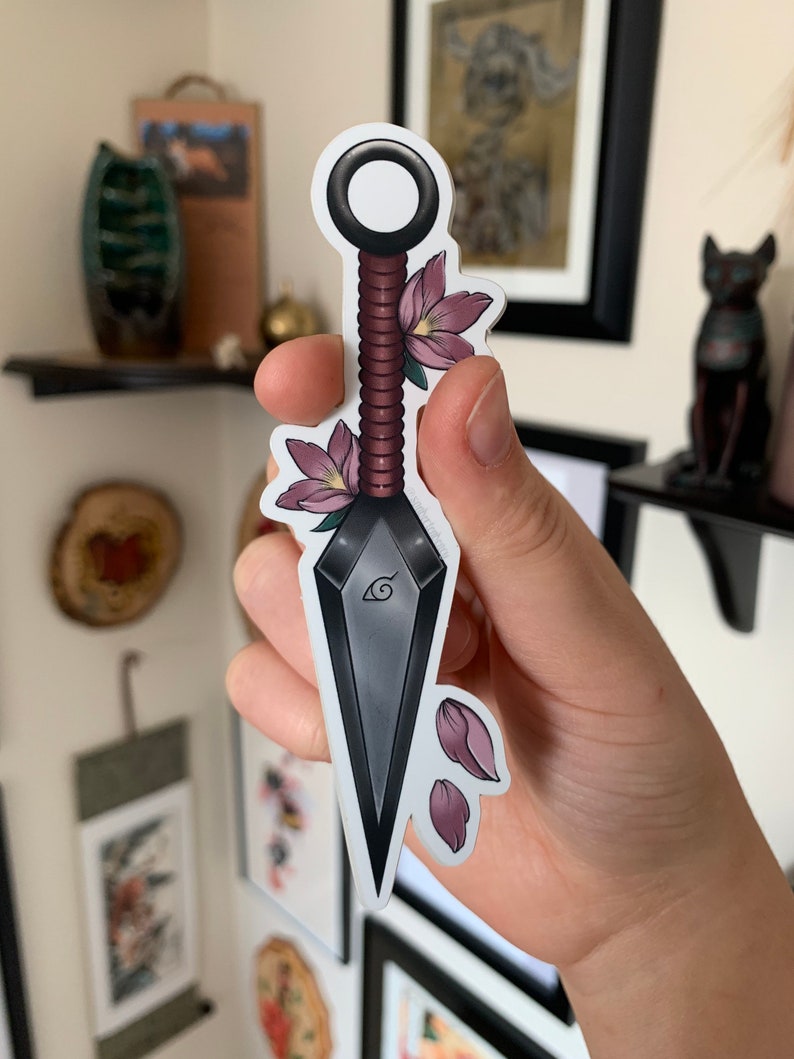 Puede incluir: Un kunai gris y negro con un mango de cuerda rojo y blanco. El kunai tiene un dise&ntilde;o de espiral estilizado y una flor rosa en el mango. El kunai est&aacute; rodeado de p&eacute;talos de flores rosas.