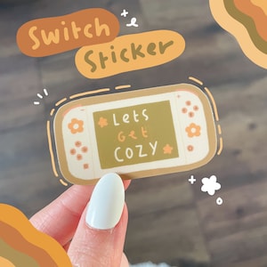 May include: A beige and brown sticker of a Nintendo Switch console with the text "Let's Get Cozy" on the screen. The sticker has a white outline and a flower design on the console.