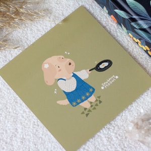 Op de afbeelding: Een groene kaart met een cartoonillustratie van een hond die een blauwe jurk draagt en een koekenpan met een ei erin vasthoudt. De tekst "Dewberry Moons" staat in wit op de kaart.