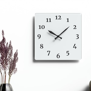 Puede incluir: Un reloj blanco con agujas negras y números negros sobre un fondo blanco. El reloj está colgado en una pared blanca. Hay una estantería de madera debajo del reloj con un jarrón de flores moradas, un oso de peluche de madera, un cuaderno y un lápiz. Hay una radio vintage en la estantería.