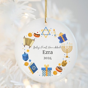 May include: White ceramic ornament with a blue and yellow design featuring a menorah, dreidel, and other Hanukkah symbols. The text "Baby's First Hanukkah Ezra 2024" is printed on the ornament.
