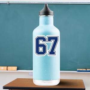 Könnte beinhalten: Eine hellblaue Wasserflasche mit schwarzem Deckel. Die Zahl "67" ist in dunkelblauer, weiß umrandeter Schrift im College-Stil dargestellt. Die Flasche steht auf einem Holzschreibtisch.
