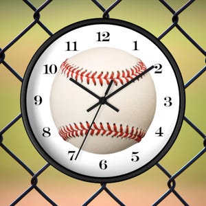 May include: A black and white baseball clock with red stitching. The clock face has black numbers and hands. The clock is hanging on a chain link fence.