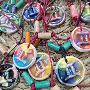 Op de afbeelding: Een verzameling kleurrijke keramische hangerkettingen. Elke ronde hanger heeft een uniek abstract ontwerp in tinten blauw, geel, oranje en paars. De kettingen hebben rode, zwarte en groene koorden met houten en cilindrische kralen.