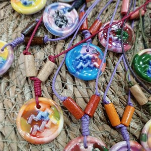 May include: A collection of colorful ceramic necklaces with zodiac symbols. Each necklace features a round pendant with a different zodiac sign painted in vibrant colors. The pendants are strung on cords with wooden beads.