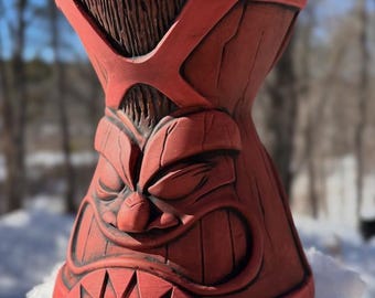 May include: A terracotta-coloured tiki mug featuring a carved face with a wide, toothy grin, narrow eyes, and a textured wood-grain pattern. The vessel has a flared top rim and a tapered base, showcasing intricate relief details in a matte finish.
