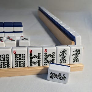 Könnte beinhalten: Ein Mahjong-Spiel mit weißen Spielsteinen und schwarzen Symbolen, darunter Zahlen und Drachen. Die Spielsteine sind auf Holzleisten mit blauen Akzenten angeordnet. Das Spielset befindet sich vor einem neutralen Hintergrund.