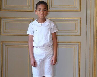 May include: A young boy wearing a white collared shirt and white shorts stands in front of a wooden door with gold trim. He is wearing black shoes.