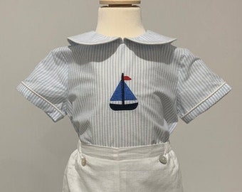 May include: A white and blue striped short-sleeved shirt with a sailor collar and a blue sailboat embroidered on the front. The shirt is paired with white linen shorts.