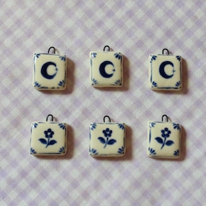 May include: Six square ceramic pendants with a blue and white floral or crescent moon design. Each pendant has a small metal loop at the top. The pendants are arranged on a light purple and white checked fabric.