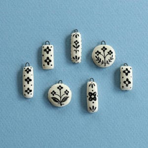 May include: A selection of white ceramic pendants featuring black floral patterns and small metal loops. The pendants are in diverse shapes, such as cylinders and circles, set against a light blue backdrop. The designs include stylized flowers and botanical motifs.