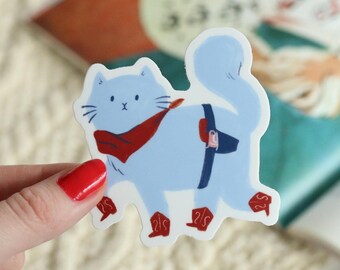 May include: A light blue cat sticker with a red scarf and boots, and a dark blue backpack. The sticker is held by a hand with red nail polish. The background includes a book with illustrations.