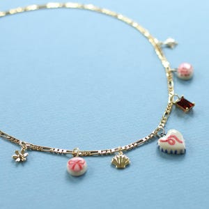 May include: Gold-coloured anklet featuring charms such as a star, a pink bead, a red rectangle, a heart, a flower, a round charm with a bow, and a seashell. The anklet is set against a light blue backdrop.