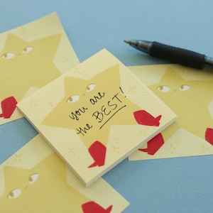 May include: A stack of square yellow sticky notes featuring a cartoon star character with eyes and red boots. The top note has the handwritten black ink text 'you are the BEST!' with small star accents. A black ballpoint pen rests nearby.