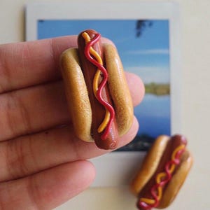 Puede incluir: Dos dijes miniatura en forma de perrito caliente hechos de arcilla polim&eacute;rica. Cada pieza presenta una salchicha marr&oacute;n en un pan beige, decorada con finas l&iacute;neas onduladas de k&eacute;tchup rojo y mostaza amarilla. El pan tiene una textura lisa.