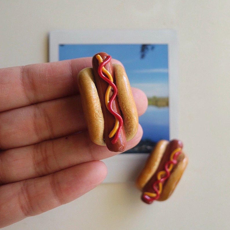 Puede incluir: Dos dijes miniatura en forma de perrito caliente hechos de arcilla polim&eacute;rica. Cada pieza presenta una salchicha marr&oacute;n en un pan beige, decorada con finas l&iacute;neas onduladas de k&eacute;tchup rojo y mostaza amarilla. El pan tiene una textura lisa.