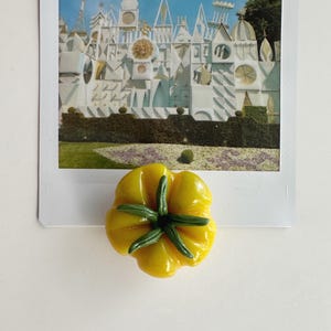 May include: A yellow, flower-shaped magnet with green accents, placed on a vintage postcard featuring a whimsical architectural scene. The magnet's design resembles a stylized flower, adding a touch of color to the image.