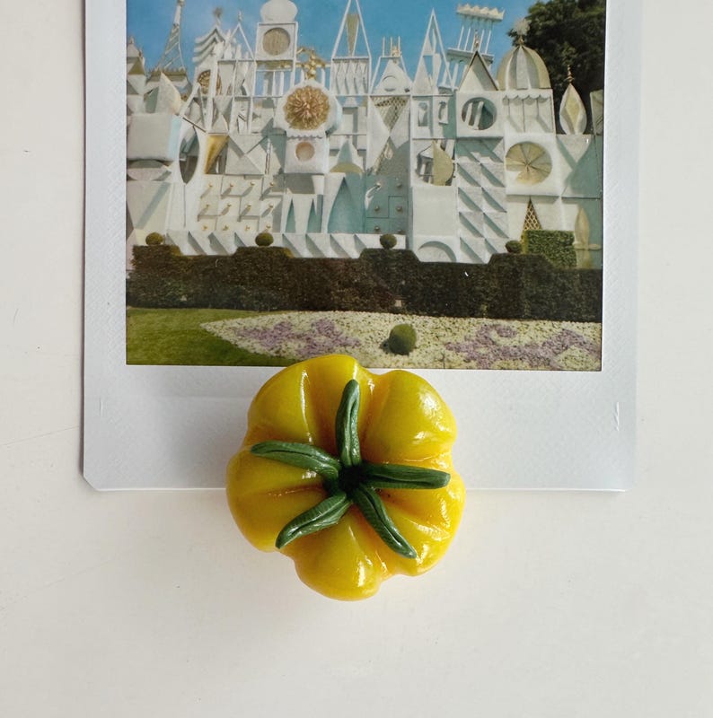 May include: A yellow, flower-shaped magnet with green accents, placed on a vintage postcard featuring a whimsical architectural scene. The magnet's design resembles a stylized flower, adding a touch of color to the image.