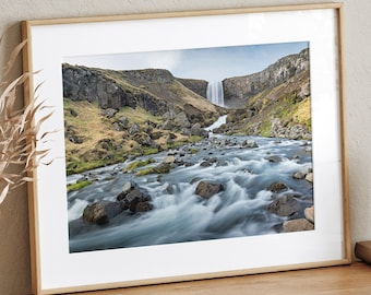 May include: A framed photograph of a waterfall cascading down a rocky cliffside into a river. The water is flowing quickly and there are rocks and greenery in the foreground.