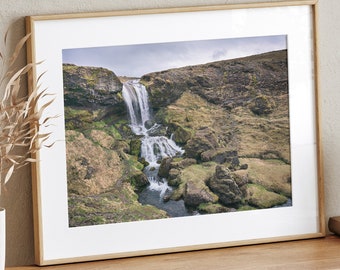 May include: A framed photograph of a waterfall cascading down a rocky hillside. The waterfall is surrounded by lush green vegetation and the sky is a cloudy gray.