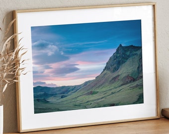 May include: A framed photograph of a mountain range with a blue sky and pink clouds. The mountain range is green and brown with a rocky peak. The photo is in a light wood frame.