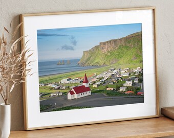 May include: A framed photograph of a white church with a red roof, situated on a green hillside overlooking a town and the ocean. The church is in the foreground, with the town and ocean in the background. The photo is framed in a light wood frame.