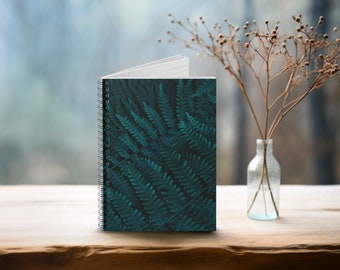 May include: A spiral bound notebook with a green fern pattern on the cover. The notebook is sitting on a wooden surface next to a glass vase with dried branches.
