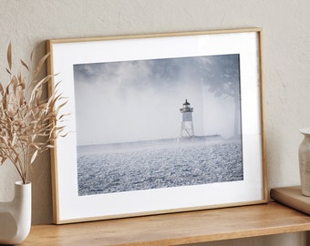 May include: A framed photograph of a lighthouse on a foggy day. The lighthouse is white with a black roof and is located on a small island in the middle of the ocean. The water is choppy and the sky is overcast.