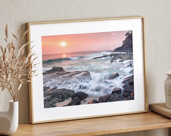 May include: A framed photograph of a seascape with a rocky shore and waves crashing against the rocks. The sky is a soft pink and orange, and the sun is setting in the distance.
