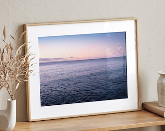 May include: A framed photograph of a seascape with a pink and purple sunset over a calm ocean. The frame is light wood and the photo is on a wooden shelf with a vase of dried flowers and other decor.