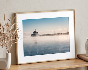 May include: A framed photograph of a lighthouse on a pier with fog rolling in over the water. The lighthouse is a white building with a red roof and is located on a pier with wooden beams. The water is calm and the fog is thick, creating a mysterious and atmospheric scene.