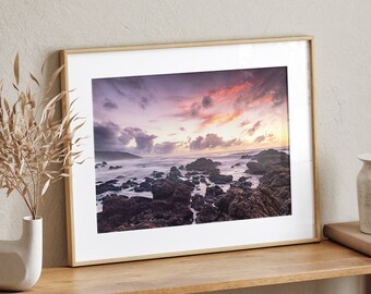 May include: A framed photograph of a rocky beach at sunset. The sky is a mix of purple, pink, and orange. The water is a deep blue and the rocks are a dark brown.