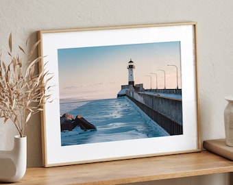 May include: A framed photograph of a lighthouse on a pier extending into a body of water. The water is partially frozen and the sky is a soft blue with a hint of pink. The lighthouse is white with a black top and a red light.