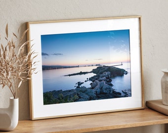 May include: A framed photograph depicting a seascape with a rocky shoreline and a distant city skyline. The sky is a soft blue with hints of pink and orange, suggesting a sunrise or sunset. The water is calm and reflective, with a slight ripple effect.