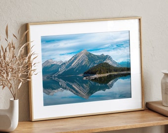 May include: A framed photograph of a mountain range reflected in a still lake. The mountains are a dark green and brown color, and the lake is a light blue. The sky is a light blue with white clouds.