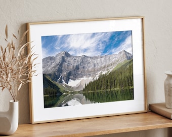 May include: A framed photograph of a mountain lake with a reflection of the surrounding mountains in the water. The lake is surrounded by trees and the mountains are snow-capped.