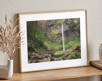 May include: A framed photograph of a waterfall cascading down a rocky cliffside in a lush green forest. The waterfall is surrounded by trees and foliage, and a small wooden bridge crosses a stream in the foreground.